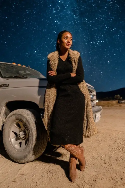 A woman wears a black long-sleeve sweater dress with a fur-trimmed coat and brown cowboy boots