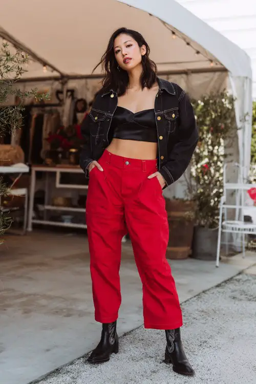A woman wears a black satin cami tucked into red high-waisted trousers, layered with a cropped denim jacket and black cowboy boots