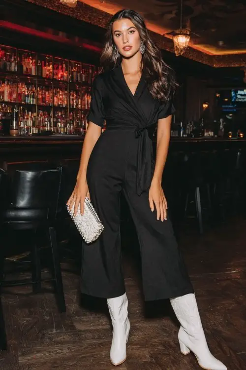 A woman wears a black wrap-front jumpsuit with a cinched waist and wide legs, finished with glossy white cowboy boots