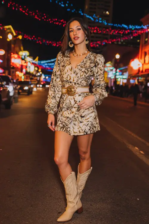 A woman wears a bold animal-print mini dress with beige cowboy boots, styled with a wide belt and statement earring