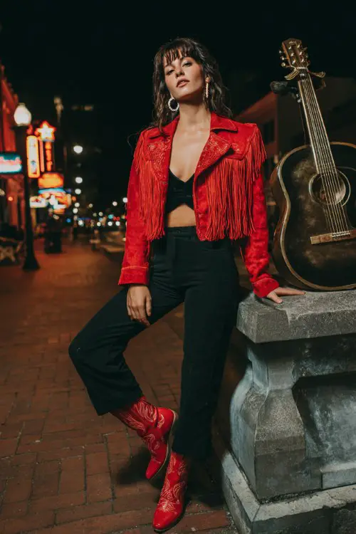 A woman wears a cropped red fringe jacket over a black fitted jumpsuit, accessorized with red cowboy boots