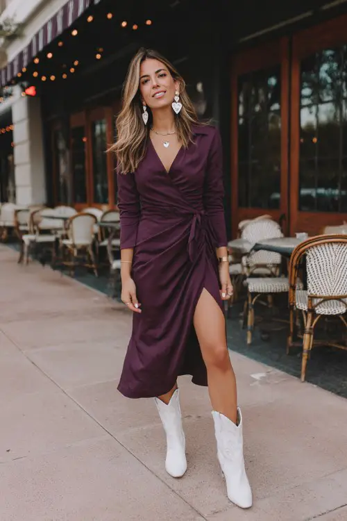 A woman wears a deep plum wrap dress with a high slit, oversized heart earrings, white cowboy boots