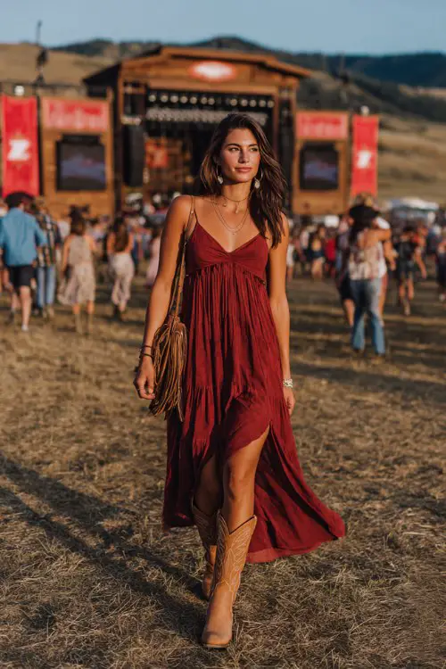 A woman wears a deep red flowy maxi dress with boho jewelry, tan western boots, and a brown fringe bag