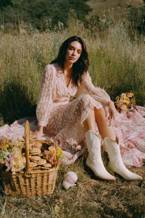 A woman wears a delicate pale blush lace dress with long sleeves and white cowgirl boots