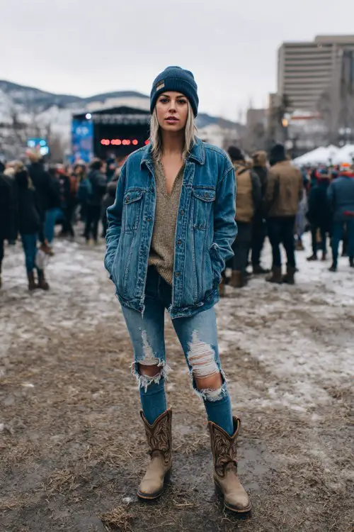 A woman wears a denim jacket layered over a thermal shirt, ripped jeans, and rugged cowboy boots