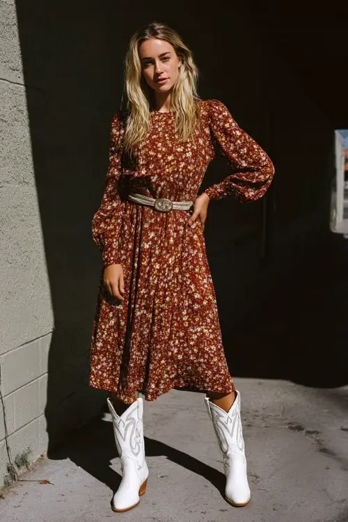 A woman wears a flowy floral midi dress with long sleeves, cinched at the waist with a slim belt, and paired with white embroidered cowboy boots