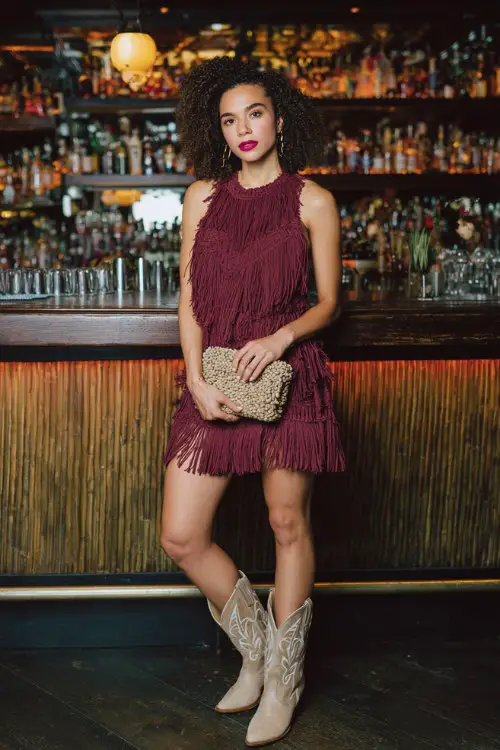 A woman wears a fringed mini dress in deep burgundy with beige cowboy boots, styled with a textured clutch and bold makeup (2)