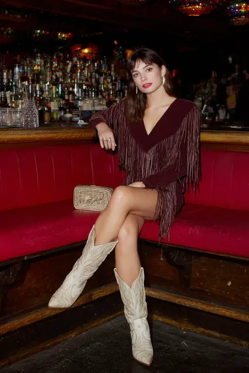 A woman wears a fringed mini dress in deep burgundy with beige cowboy boots, styled with a textured clutch and bold makeup