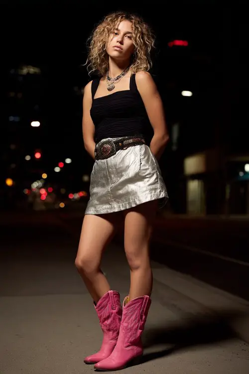 A woman wears a metallic silver mini skirt with a fitted black top and bright pink cowboy boots, paired with a statement belt and layered jewelry