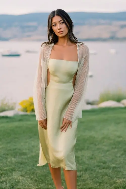 A woman wears a minimalist silk slip dress layered with a cropped organza jacket and strappy heels, lakeside spring wedding setting, soft wind movement