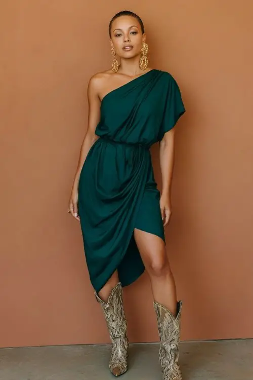 A woman wears a one-shoulder wrap dress in deep emerald, styled with snakeskin cowboy boots and bold gold earrings