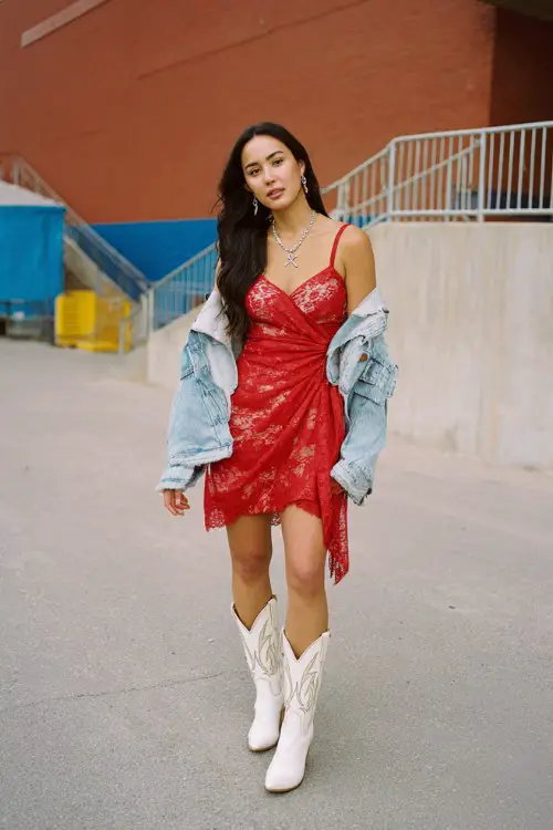 A woman wears a red lace asymmetrical dress with a denim jacket and white cowboy boots