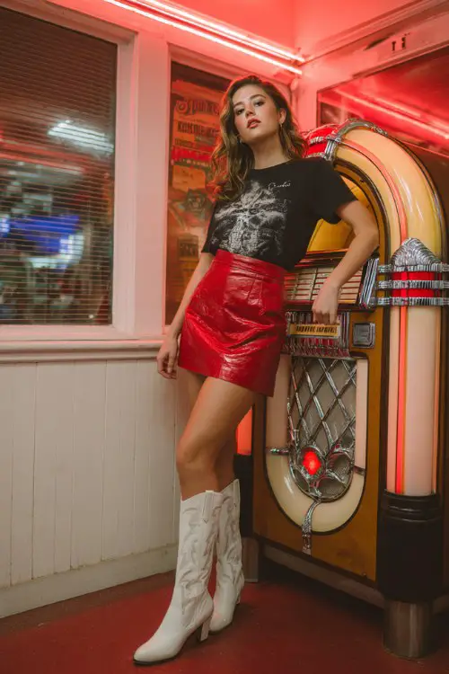 A woman wears a red leather mini skirt with a black graphic tee and white cowboy boots