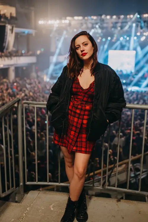 A woman wears a red plaid mini dress under a cropped black bomber jacket, ankle boots, and bold red lipstick