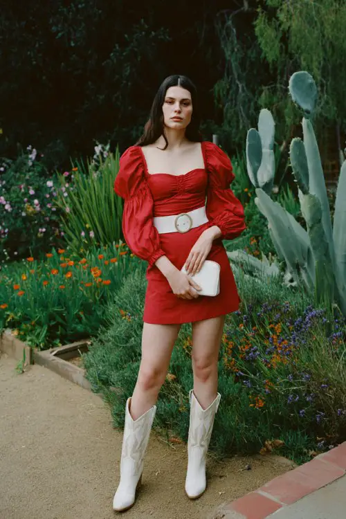 A woman wears a red puff-sleeve mini dress with a white waist belt and white cowboy boots