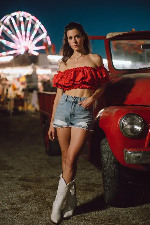 A woman wears a ruffled off-the-shoulder red crop top with high-waisted denim shorts and white cowboy boots