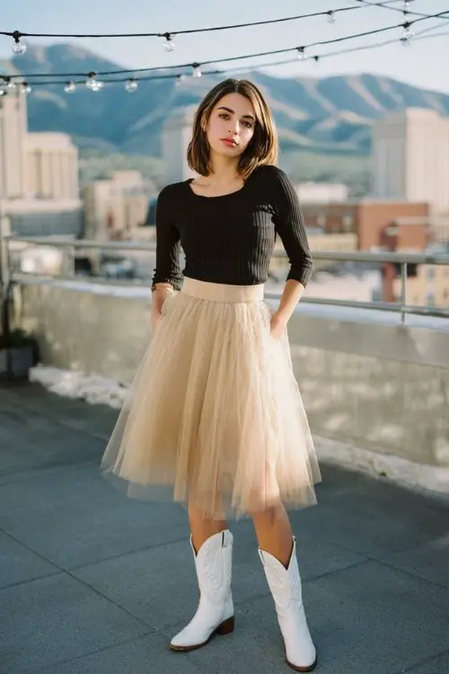A woman wears a soft beige tulle skirt with a fitted black ribbed top and white cowboy boots, standing under string lights