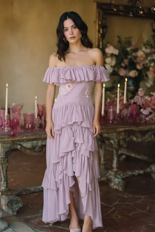 A woman wears a soft lilac off-shoulder dress with ruffled tiers and embroidered roses, paired with blush heels