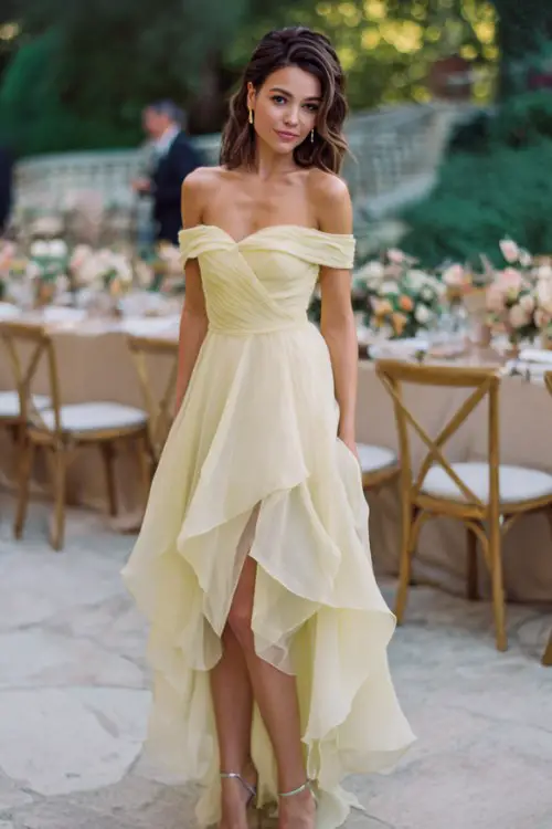 A woman wears a sophisticated off-shoulder gown in pale yellow chiffon with layered skirt movement, metallic sandals adding sparkle