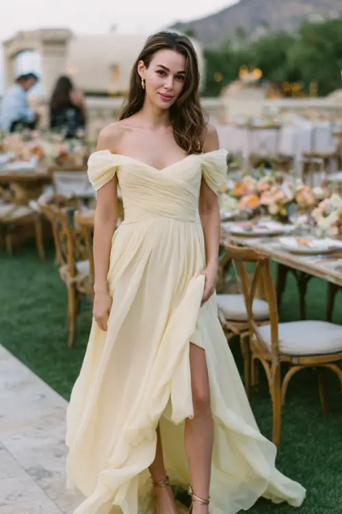 A woman wears a sophisticated off-shoulder gown in pale yellow chiffon with layered skirt movement, metallic sandals adding sparkle