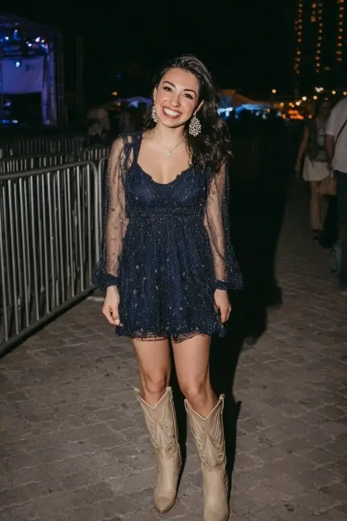 A woman wears a sparkly navy A-line mini dress with sheer sleeves, styled with beige cowboy boots and chandelier earrings