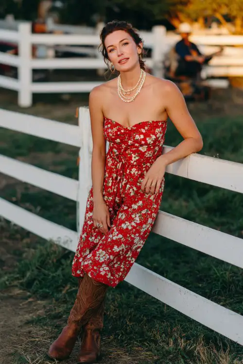 A woman wears a strapless red floral romper styled with layered pearl necklaces and brown tall boots