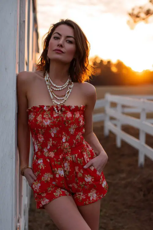 A woman wears a strapless red floral romper styled with layered pearl necklaces and brown tall boots