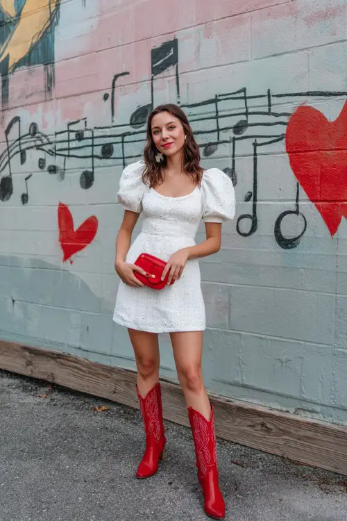 A woman wears a white eyelet mini dress with puff sleeves, red cowboy boots, and a matching red clutch