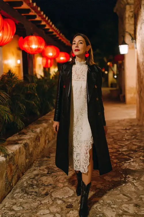 A woman wears a white lace dress under a longline black coat with black cowboy boots and red heart earrings