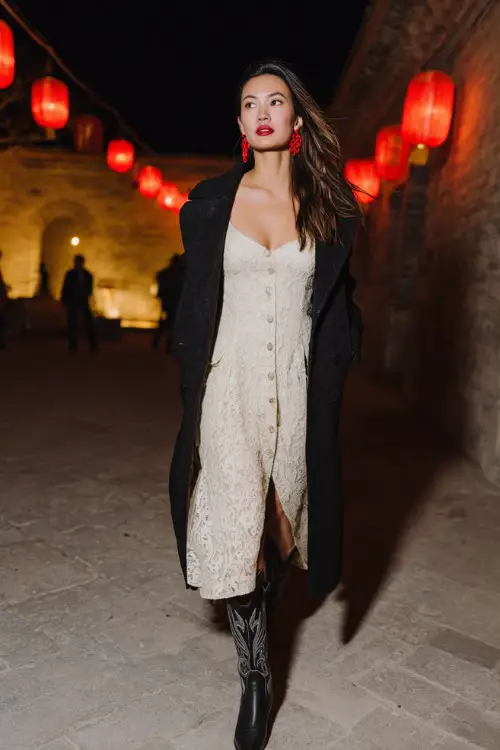 A woman wears a white lace dress under a longline black coat with black cowboy boots and red heart earrings