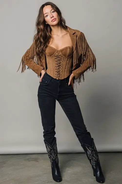 A woman wears dark denim skinny jeans paired with a structured corset top and a cropped suede fringe jacket