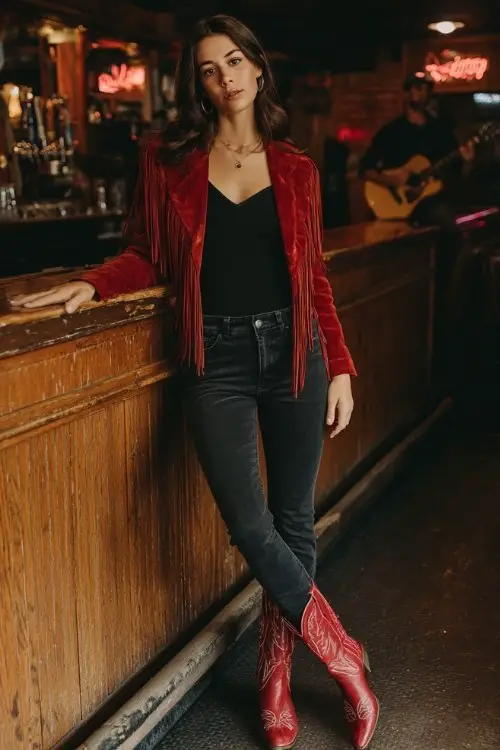 A woman wears dark skinny jeans with a body-hugging black top and a red suede fringe jacket, styled with classic red cowboy boots