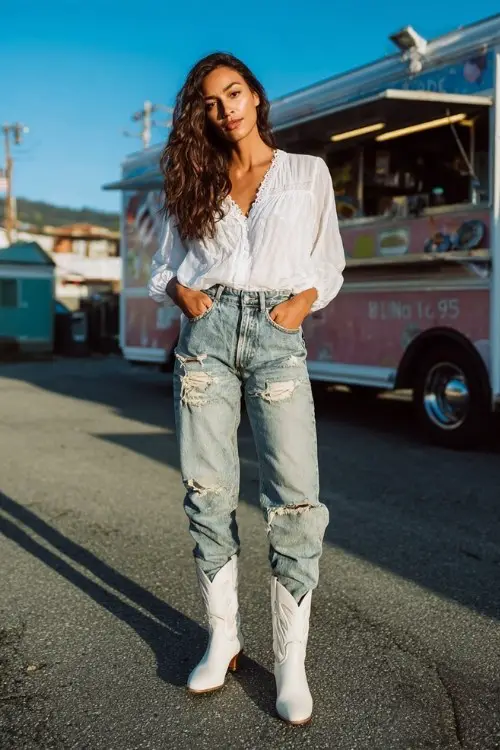 A woman wears distressed jeans, a flowy white blouse, and white cowboy boots