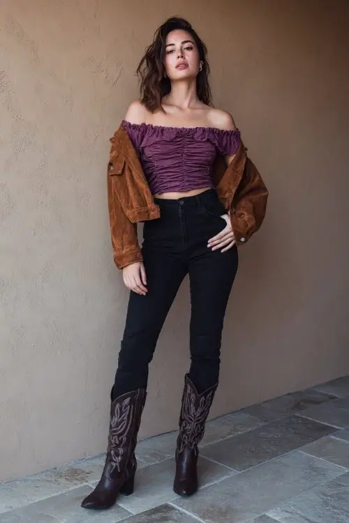A woman wears high-waisted black jeans paired with a ruched off-shoulder top in plum, layered under a cropped suede jacket and styled with classic western boots