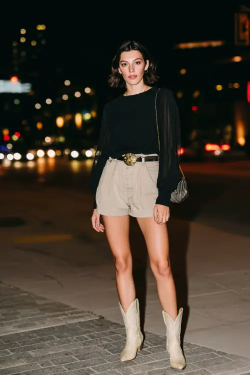 A woman wears high-waisted black shorts with a sheer long-sleeve blouse and beige cowboy boots