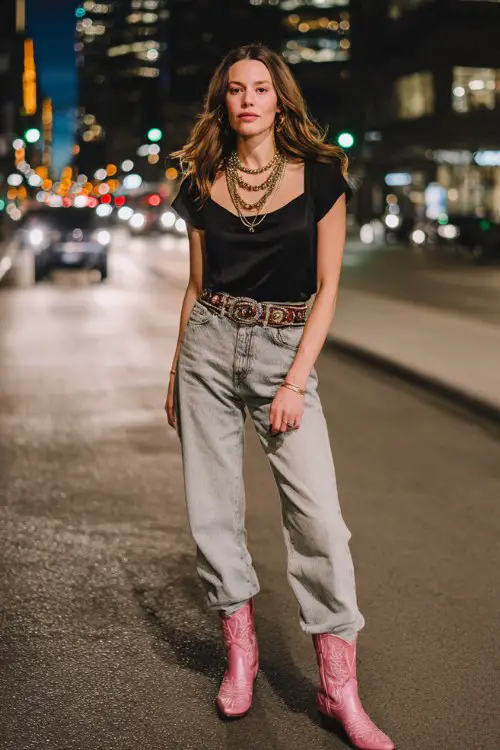 A woman wears light-wash straight-leg jeans with a silky black cami and pink cowboy boots, paired with a statement belt and layered necklaces