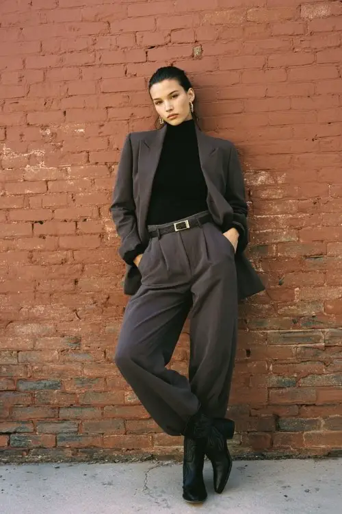 A woman wears wide-leg trousers with a fitted turtleneck and a belted long blazer, finished with black cowboy boots