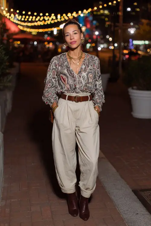 a woman over 40 wears a flowy patterned blouse tucked into high-waisted neutral pants with a leather belt and ankle boots, accessorized with layered necklaces