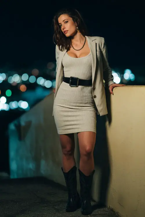 A curvy woman wears a bodycon knit dress styled with black cowboy boots, layered with an oversized blazer and statement belt accentuating the waist