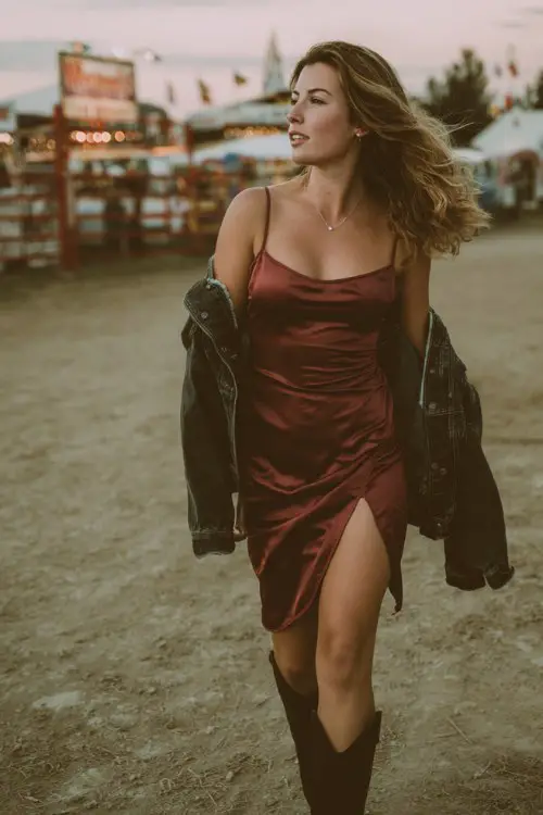 A curvy woman wears a burgundy slip dress with a side slit and black cowboy boots, styled with a black denim jacket draped over one shoulder