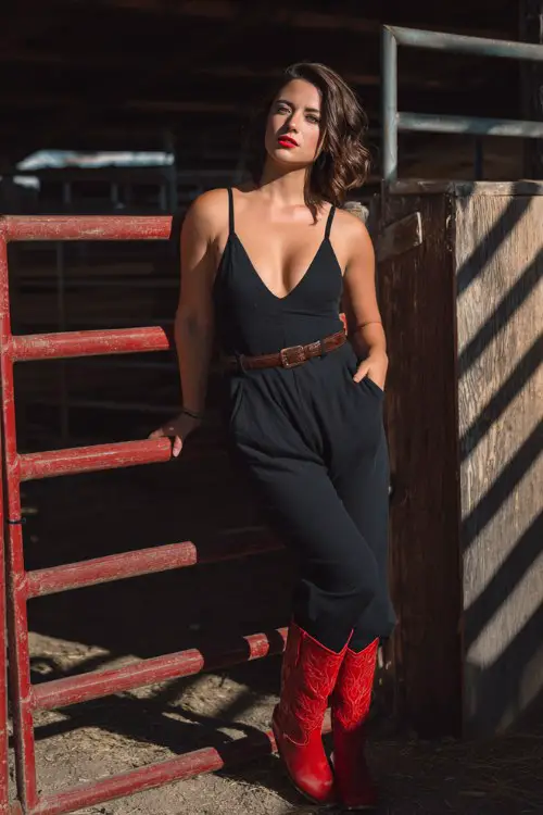 A curvy woman wears a fitted black jumpsuit with a deep V-neck, cinched with a leather belt, and bright red cowboy boots