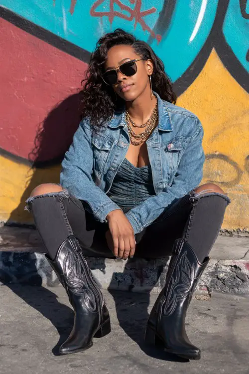 A curvy woman wears distressed high-waisted jeans tucked into glossy black cowboy boots, paired with a fitted bodysuit and oversized denim jacket