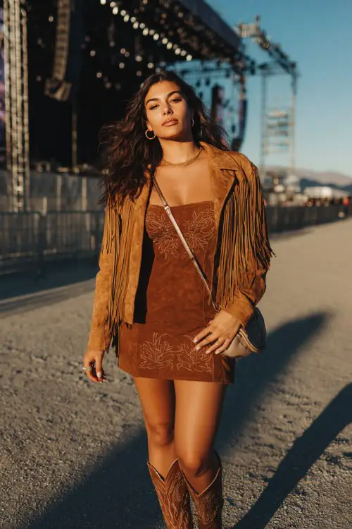 A curvy woman wears embroidered brown cowboy boots with a bodycon mini dress and fringe suede jacket, accessorized with statement rings and crossbody bag