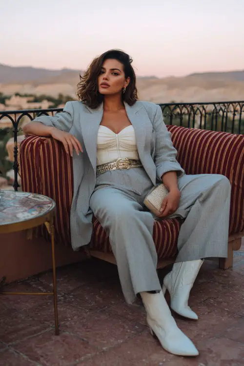 A curvy woman wears wide-leg tailored trousers styled with white cowboy boots, a corset-inspired top and oversized blazer, accessorized with a statement belt and clutch