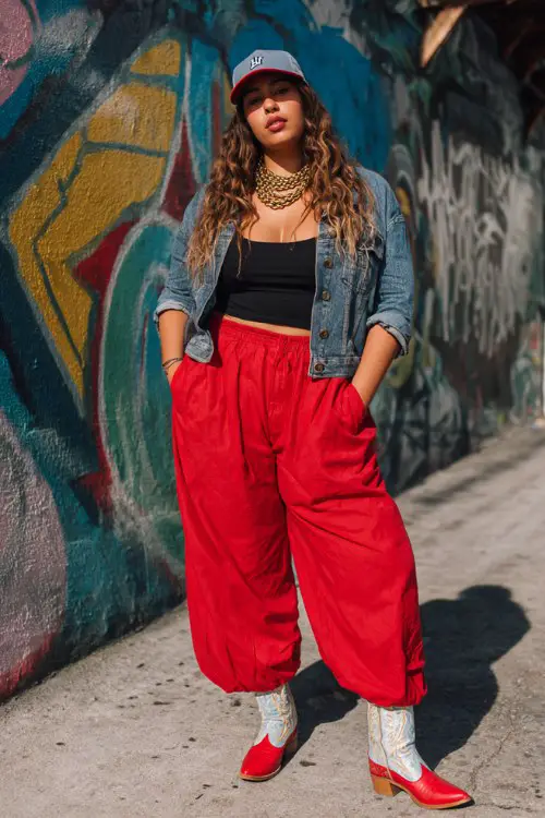 A curvy woman wears wide-leg trousers with red cowboy boots, a fitted bodysuit and cropped denim jacket, accessorized with layered chains and a baseball cap
