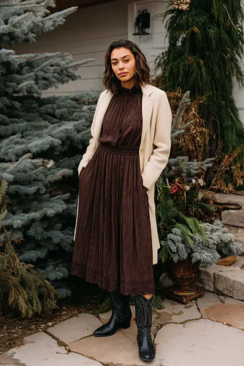 A full-body image of a woman in a chocolate brown high-neck midi dress with pleated skirt and lace trim, styled with black cowboy boots