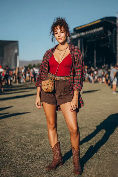 A full body woman wears brown cowboy boots with high-waisted shorts, a fitted bold red bodysuit and oversized plaid shirt worn open
