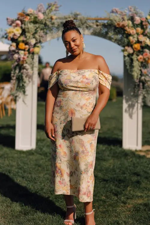 A plus-size woman over 50 wears a floral midi dress with a fitted bodice and flowy skirt, styled with block-heel sandals and a neutral clutch