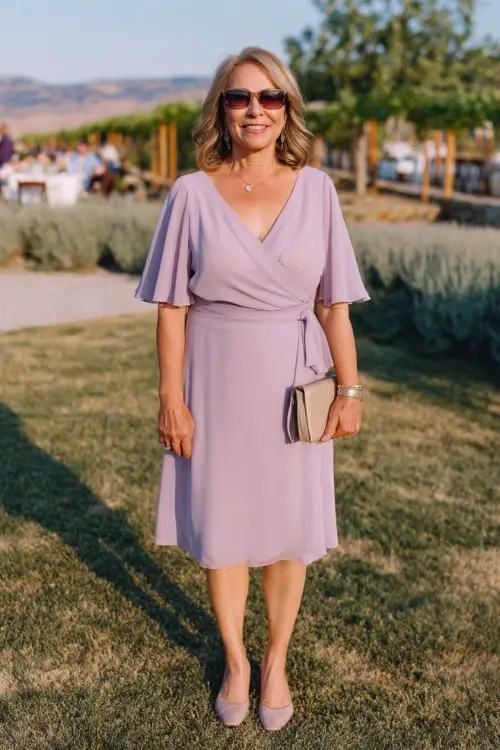 A plus-size woman over 50 wears an A-line chiffon dress in soft lavender with flutter sleeves, styled with comfortable low heels and a clutch