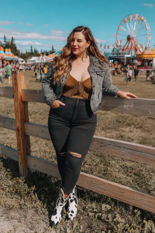 A plus-size woman wears high-rise distressed black jeans with a tucked-in fringed suede crop top, layered with a studded denim jacket and cow print cowboy boots